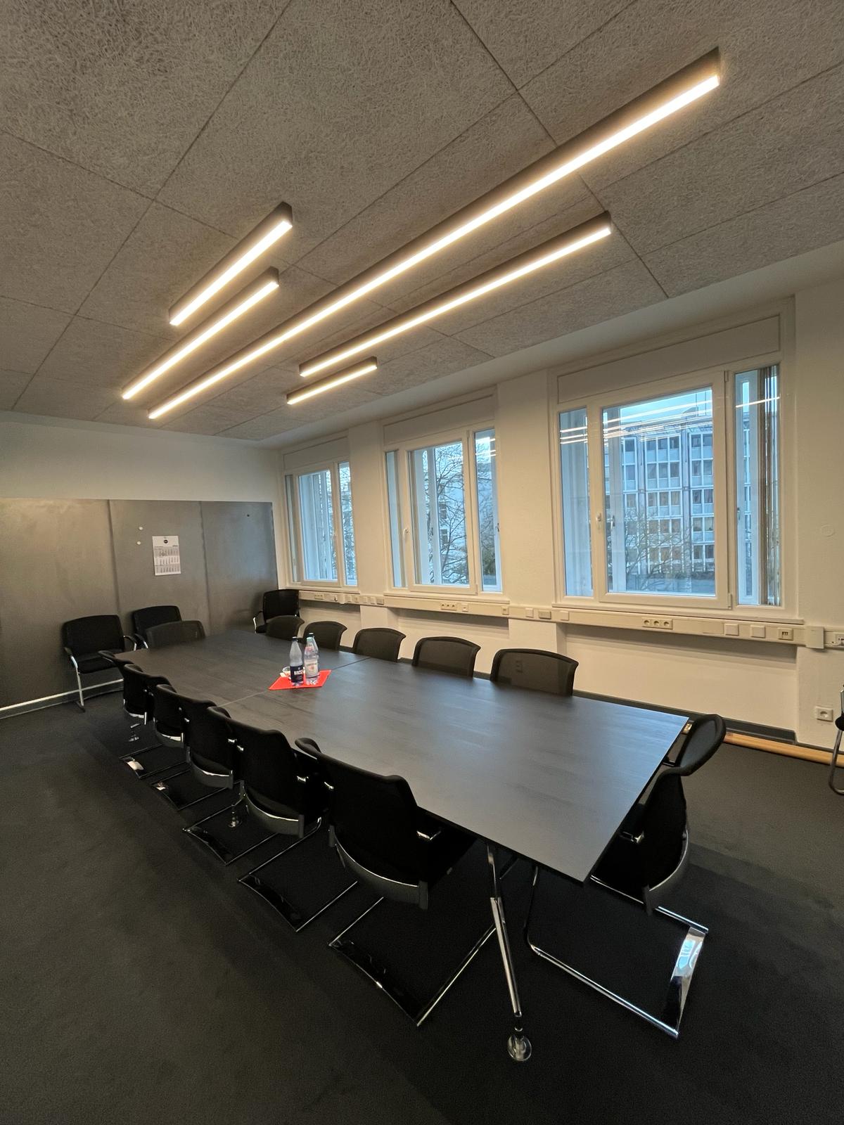 Moderne Besprechungsraum mit langen Leuchten und einem großen Tisch, umgeben von Stühlen.