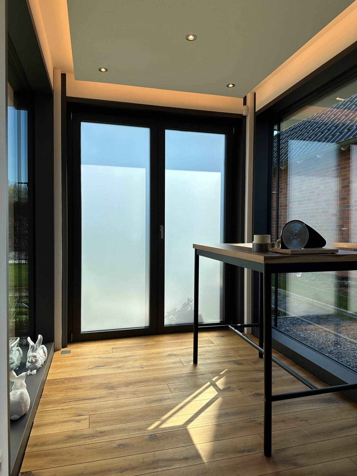 Heller Raum mit großen Fenstern, Holzfußboden und einem Tisch im Vordergrund.