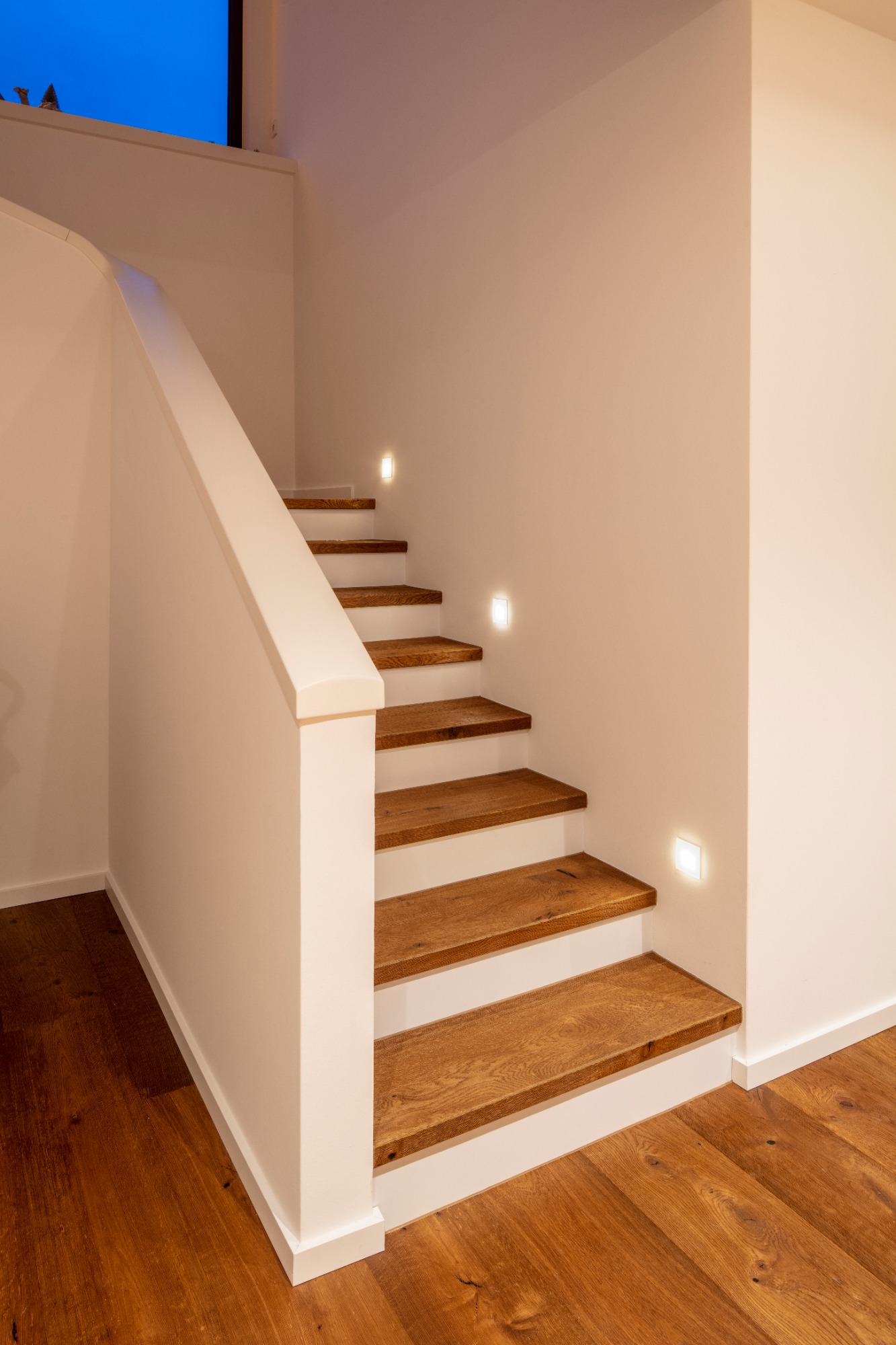 Treppenhaus von Einfamilienhaus Werne Holztreppe mit sanfter Beleuchtung, die an einer weißen Wand entlangführt.