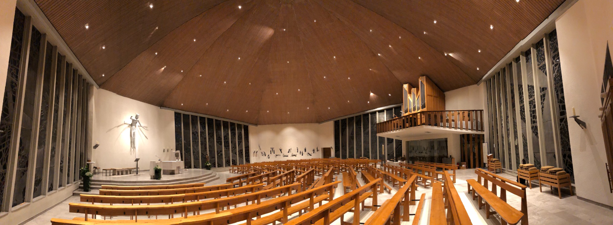 Moderne Kirche mit Holzdecke, Bänken und großer Fensterfront.