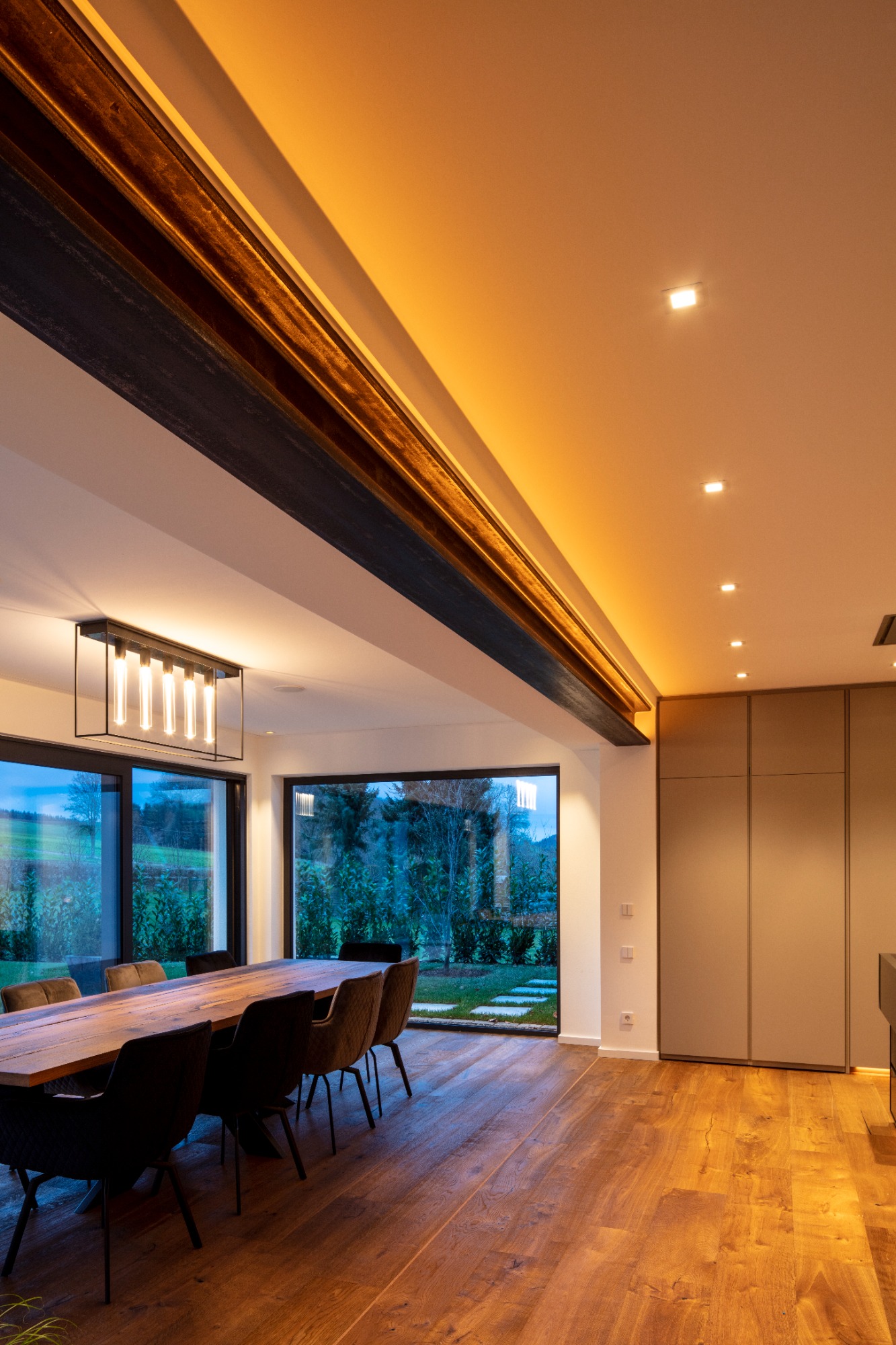 Modernes Esszimmer mit Holztisch, Stühlen und indirekter Beleuchtung, große Fenster am Abend.